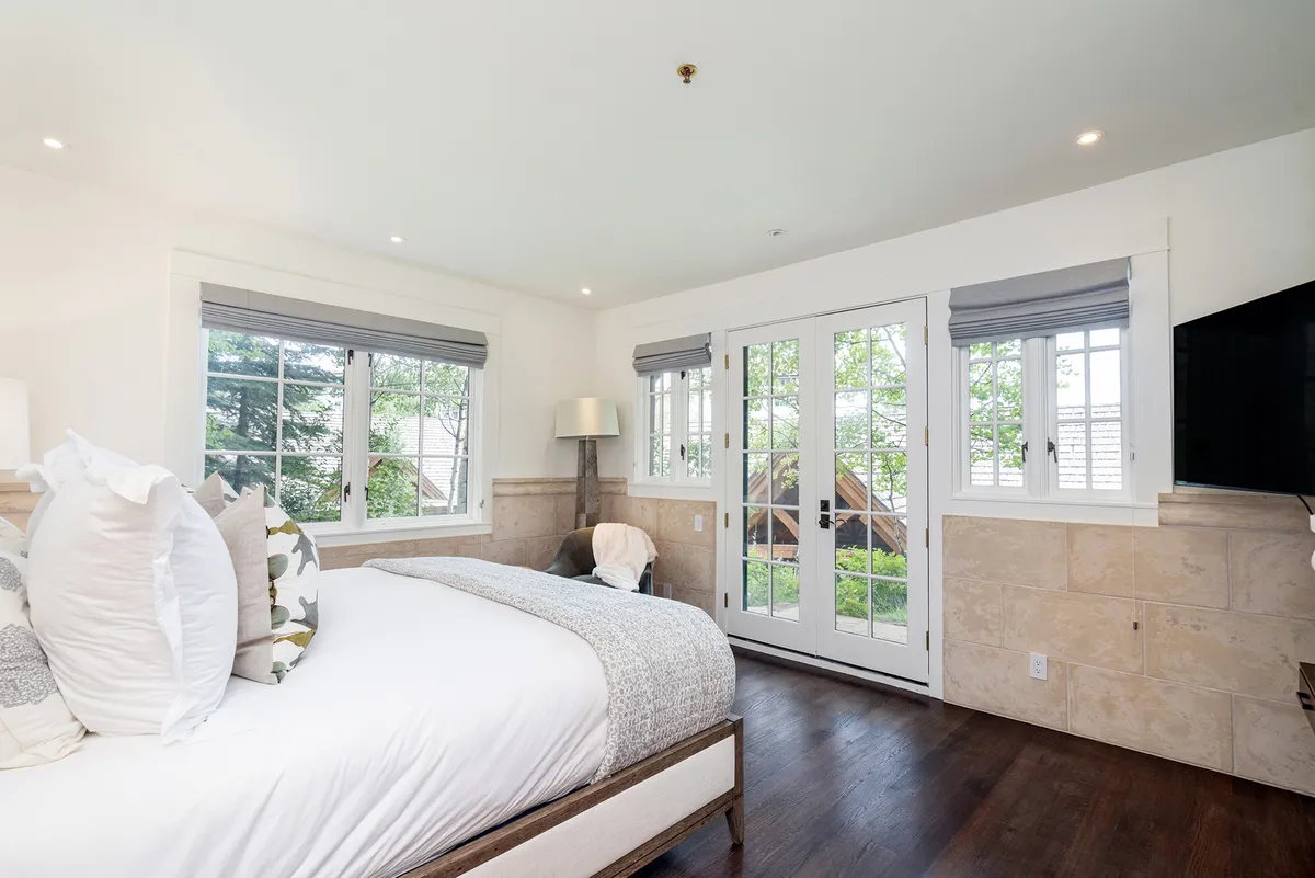 A spacious bedroom featuring a large, inviting bed adorned with decorative pillows, double doors that open to an outdoor space, a stylish lamp, and a wall-mounted TV. The room is bathed in natural light, suggesting a bright and serene atmosphere ideal for rest and relaxation.
