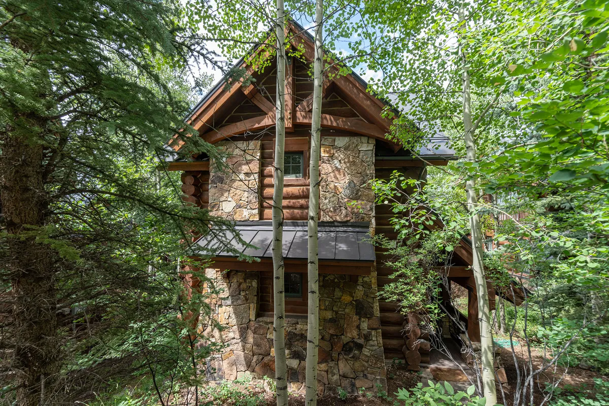 A serene exterior view of a stand-alone mountain cabin, featuring stone and wooden log construction nestled among tall trees. The cabin’s porch offers inviting spots to relax, with sunlight filtering through the forest and views of nature all around.