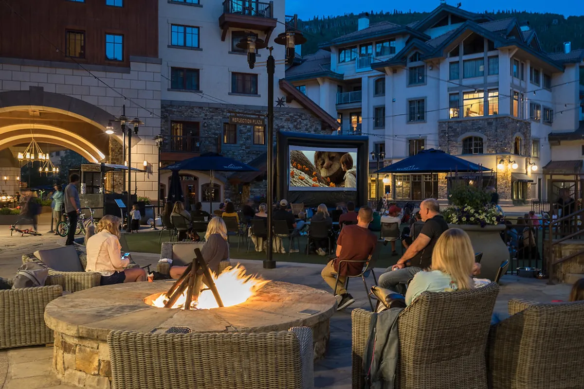 This image showcases a cozy outdoor fire pit area with comfortable seating, an outdoor movie screen, and the architectural appeal of surrounding cabins. The evening ambiance highlights relaxation and communal enjoyment under the stars, evoking mountain tranquility and the pleasures of outdoor living.
