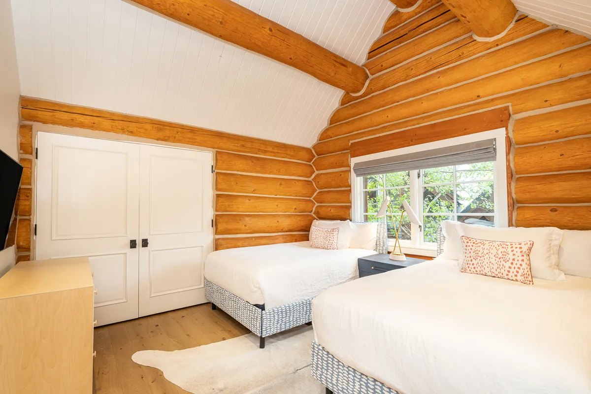 A charming bedroom with log walls, twin beds, windows that let in natural light, and a nightstand between the beds. The space feels comfortable and serene, ideal for children or extra guests to unwind and share cozy moments.