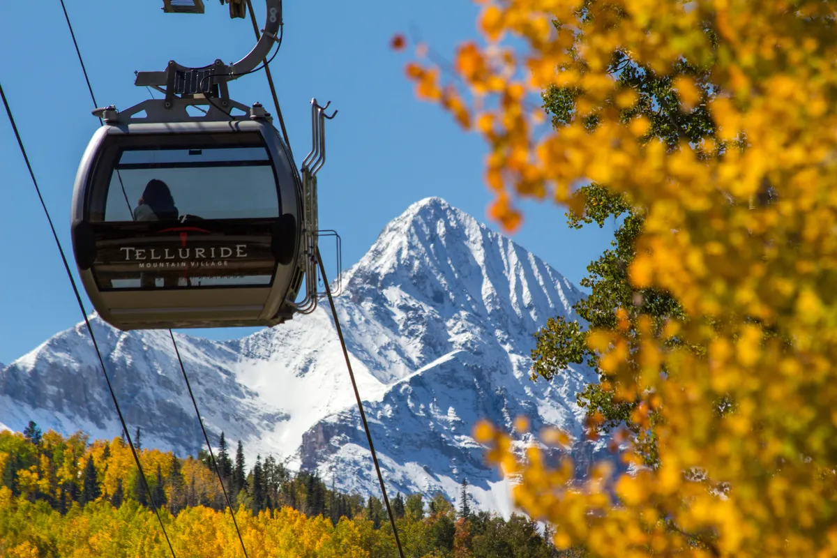 A scenic gondola gliding above colorful autumn foliage, with snow-capped mountains rising in the background. The gondola is a signature feature connecting Mountain Village to historic Telluride and offers an iconic visual link to the adventure and convenience described in the narrative.