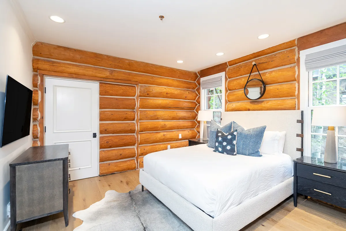 A tranquil bedroom with log walls, a comfortable bed, nightstand, decorative pillows, and a mirror. The space is filled with natural light, projecting a sense of warmth and coziness for restful family evenings or nap times for little ones.