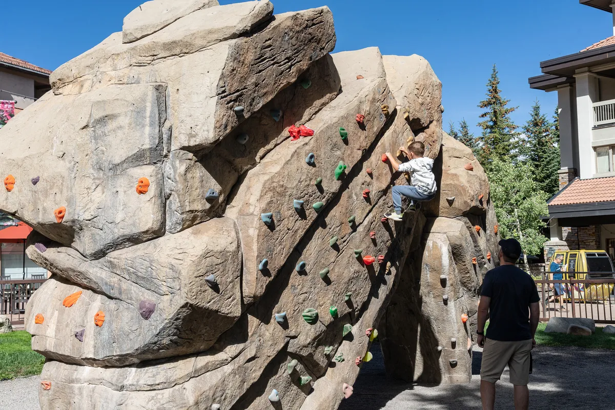 A child climbs a rock wall while an adult, likely a family member, observes encouragingly. The scene is set outdoors with green grass and blue sky, illustrating active family play and achievement in a safe, inviting environment.