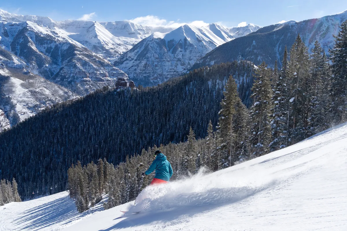 A skier rushes down a powdery slope surrounded by snow-covered mountains and dense pine trees, with the panorama capturing both action and the grandeur of the alpine landscape. The crisp air and thrilling atmosphere are palpable.