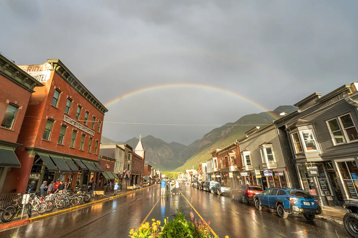 A lively village street scene featuring historic buildings, a rainbow arcing overhead, wet pavement from a recent shower, and people strolling or biking along the sidewalk. The shopfronts and café tables invite exploration and socializing, evoking the vibrant community atmosphere described in the section.