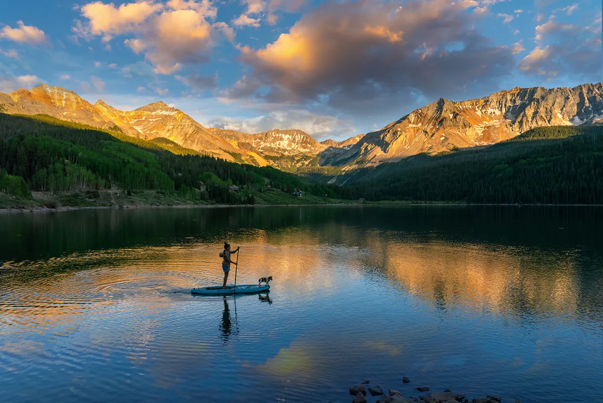 A paddleboarder and their dog glide across a reflective mountain lake in summer, with lush greenery and towering peaks in the background. The scene is tranquil yet invigorating, capturing the essence of outdoor summer exploration.