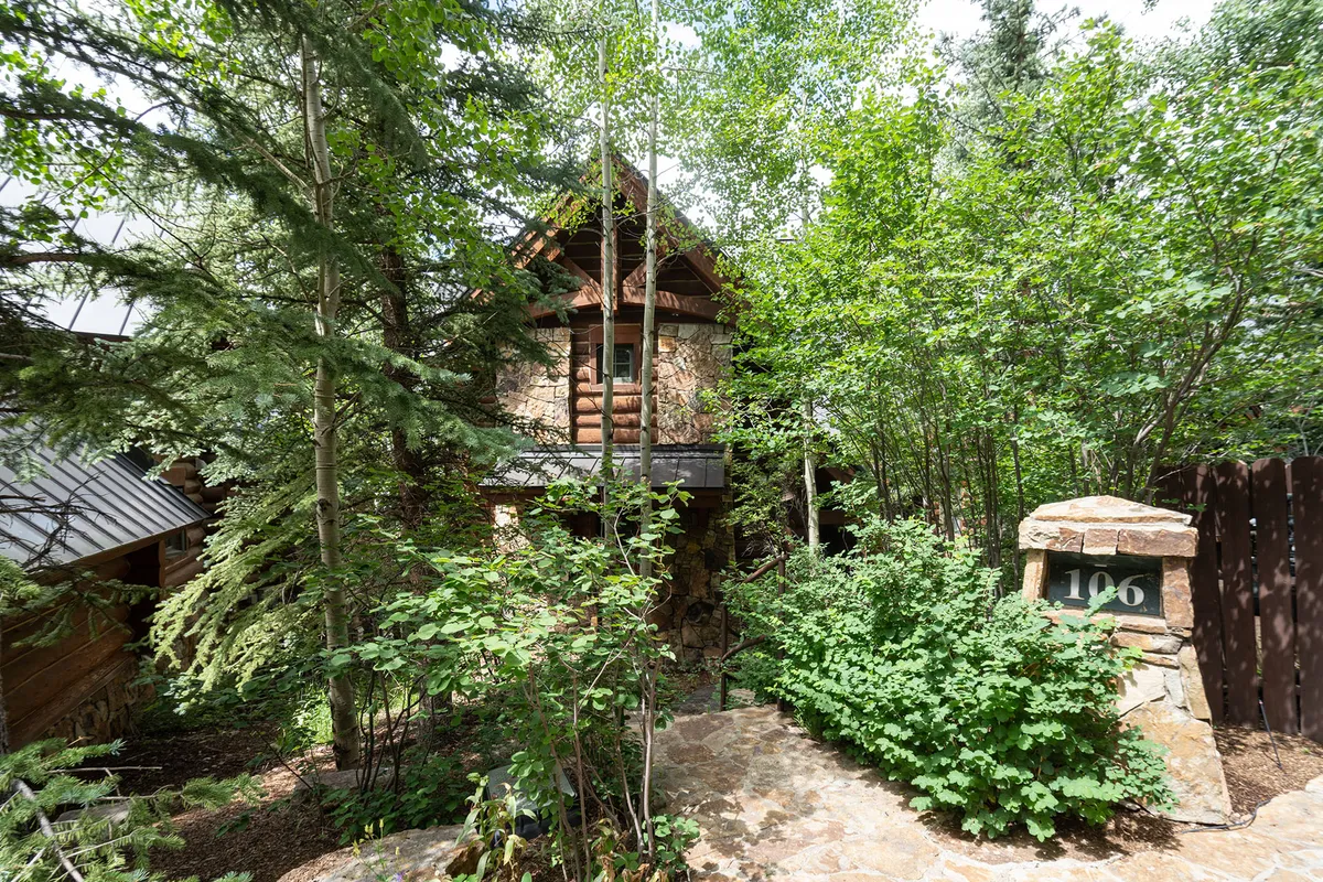 A welcoming rustic log cabin framed by lush greenery, a stone pathway leading up to the entrance, and a cozy porch area that hints at relaxing mornings or evenings outdoors. The setting feels tranquil yet connected to nature, with subtle design cues suggesting access to trails and the surrounding village.