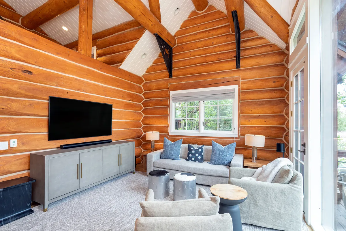 The image showcases a warmly lit living room with rustic log walls, a plush sofa adorned with cushions, a sleek large television, and a welcoming coffee table, creating a cozy yet stylish mountain retreat atmosphere.