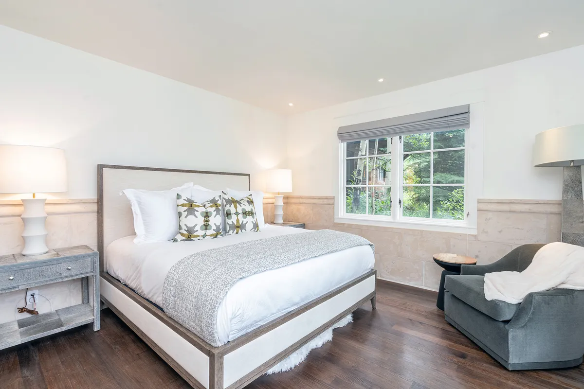 A cozy bedroom with a bed topped by decorative pillows, a comfortable armchair by a large window streaming with natural light, and stylish bedside lamps. The atmosphere is warm and inviting, perfect for relaxation or enjoying a quiet morning coffee.