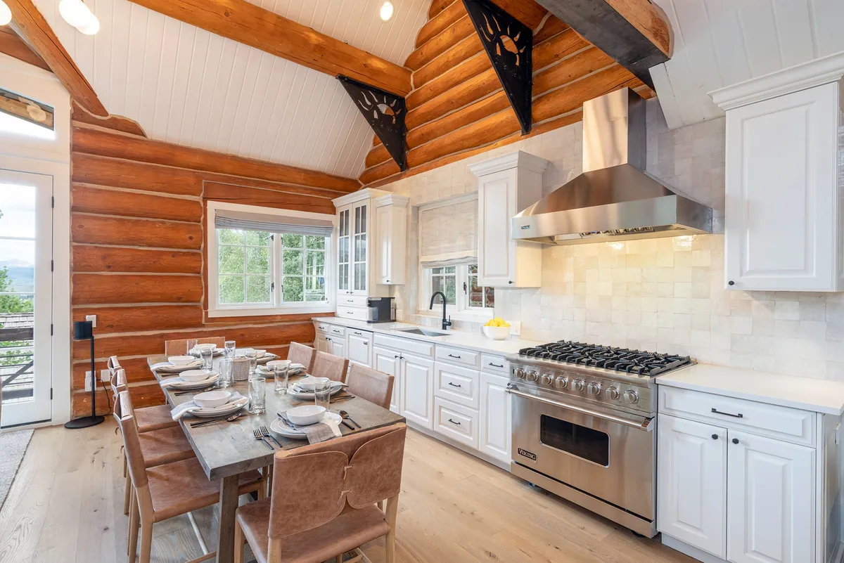This image captures a cozy and spacious interior with a large dining table set against log walls, stainless steel appliances, and wooden beams. The open-concept layout allows clear sightlines and easy movement, making it ideal for families who want to supervise children while preparing meals or enjoying time together.