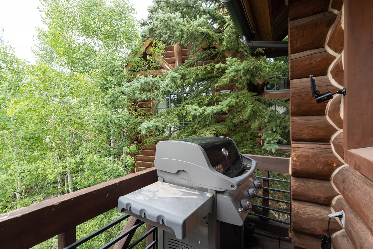 A private balcony with a grill overlooks lush greenery, featuring a log cabin structure and inviting outdoor space for al fresco dining or evening gatherings. The tranquil setting offers a seamless extension of the interior's charm to the outdoors, perfect for relaxing or entertaining under the sky.