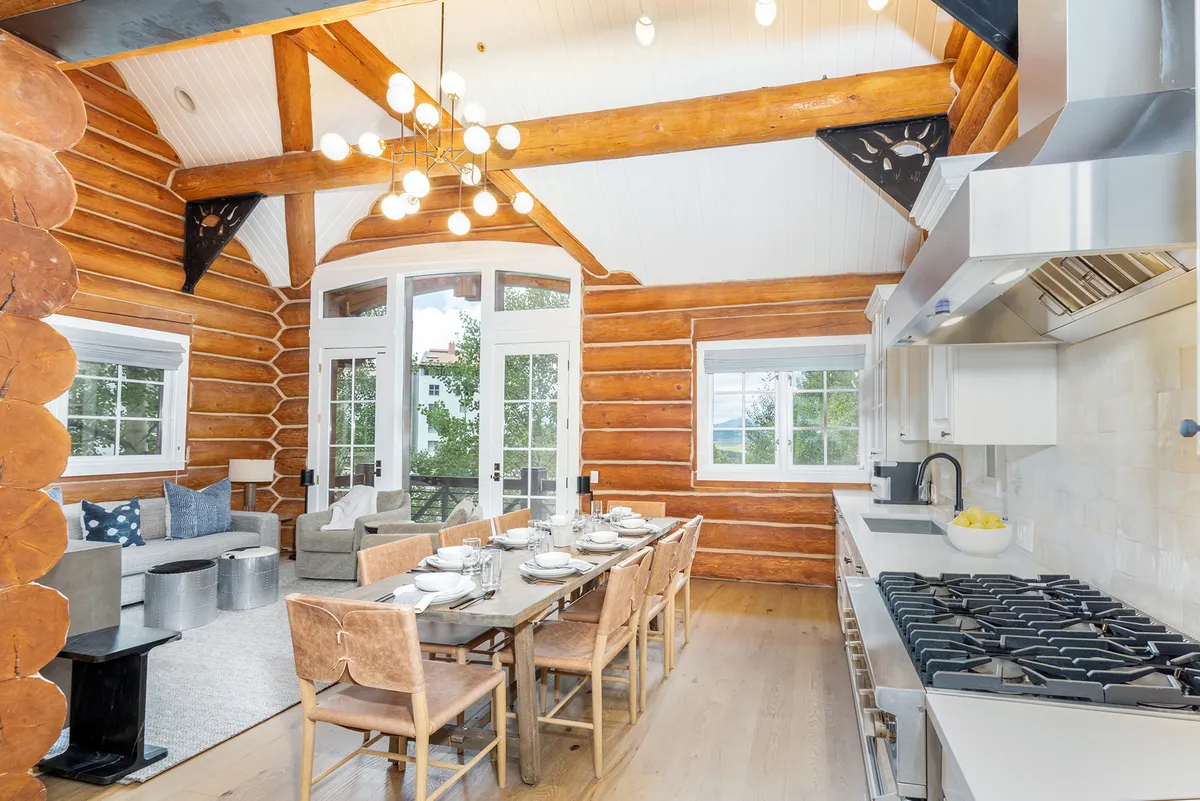 A sunlit great room featuring exposed wooden beams, a large dining table set for gathering, a spacious kitchen island, and expansive windows that flood the space with natural light. The open layout seamlessly connects the living, dining, and kitchen areas, creating a central hub for family and friends to relax, dine, and socialize.
