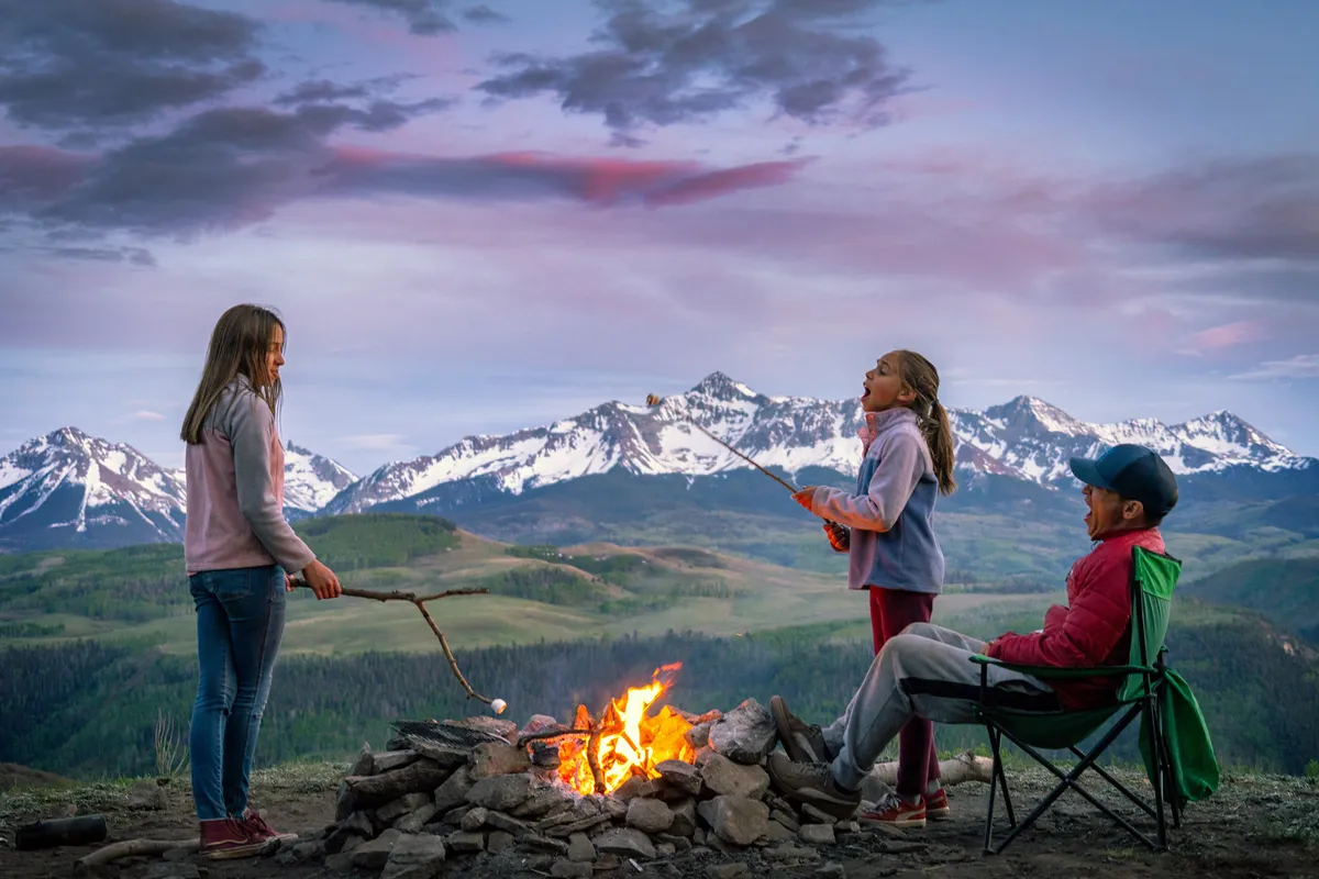 A family enjoys an evening campfire with children roasting marshmallows against a stunning mountain backdrop. Camping chairs are scattered around the fire, and the warm glow of the flames contrasts with the cool tones of the mountain scenery.