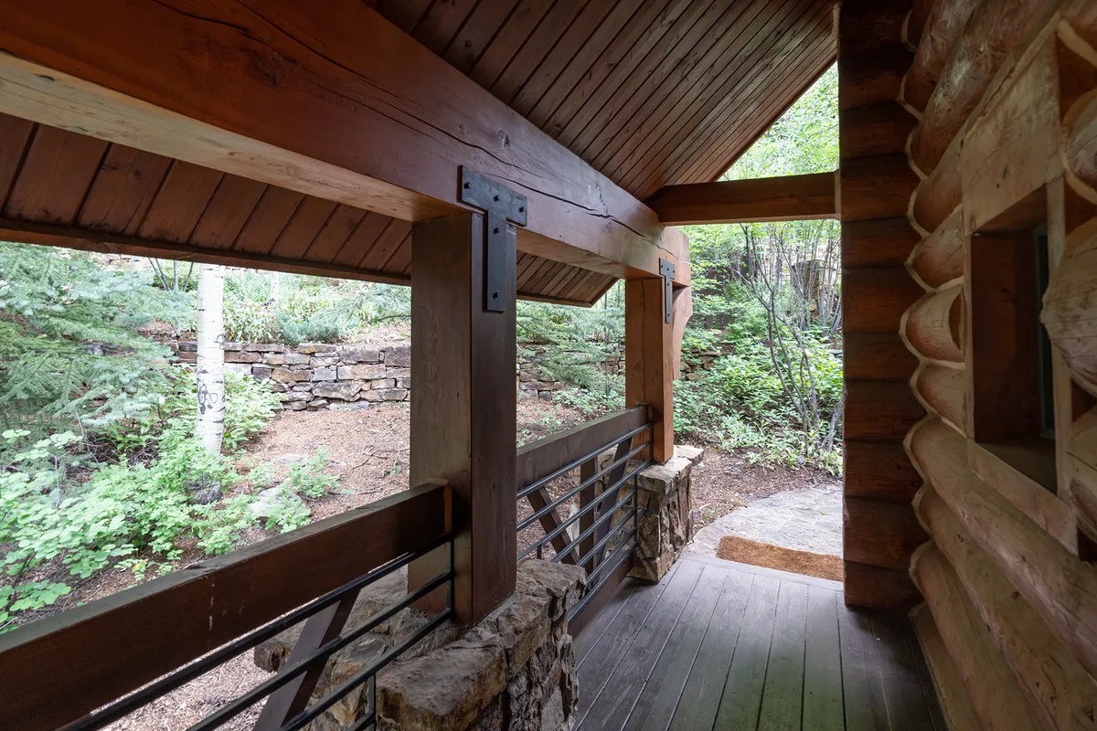 The image presents a tranquil covered porch with wooden beams and stone columns, looking out toward the serene outdoors. The scene invites viewers to imagine relaxing on the porch, immersing in a peaceful natural setting, and enjoying views of the surrounding mountains and trees.