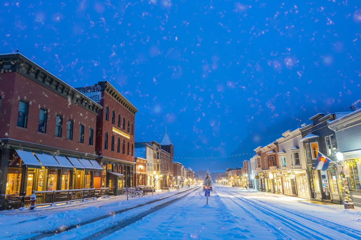 A snow-covered street lined with warmly lit storefronts and a Christmas tree glowing in the distance, evoking the atmosphere of a cozy, bustling winter village. Shoppers stroll beneath twinkling holiday lights, and the scene is framed by a picturesque wintry backdrop.